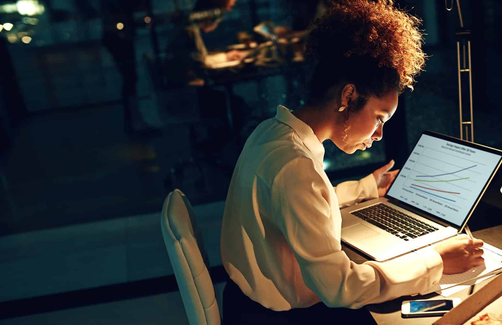 A busy woman writing down notes beside a laptop.